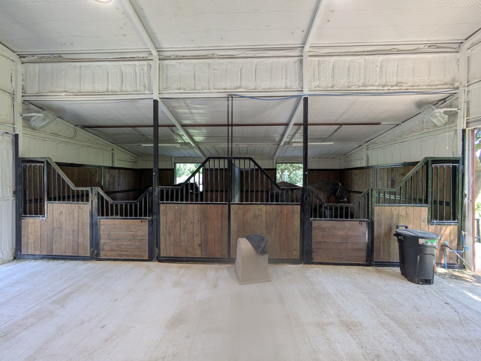 Finished row of new stalls in a Dallas-area barn: black steel fronts with stepped yokes over stained wood panels, on a concrete floor