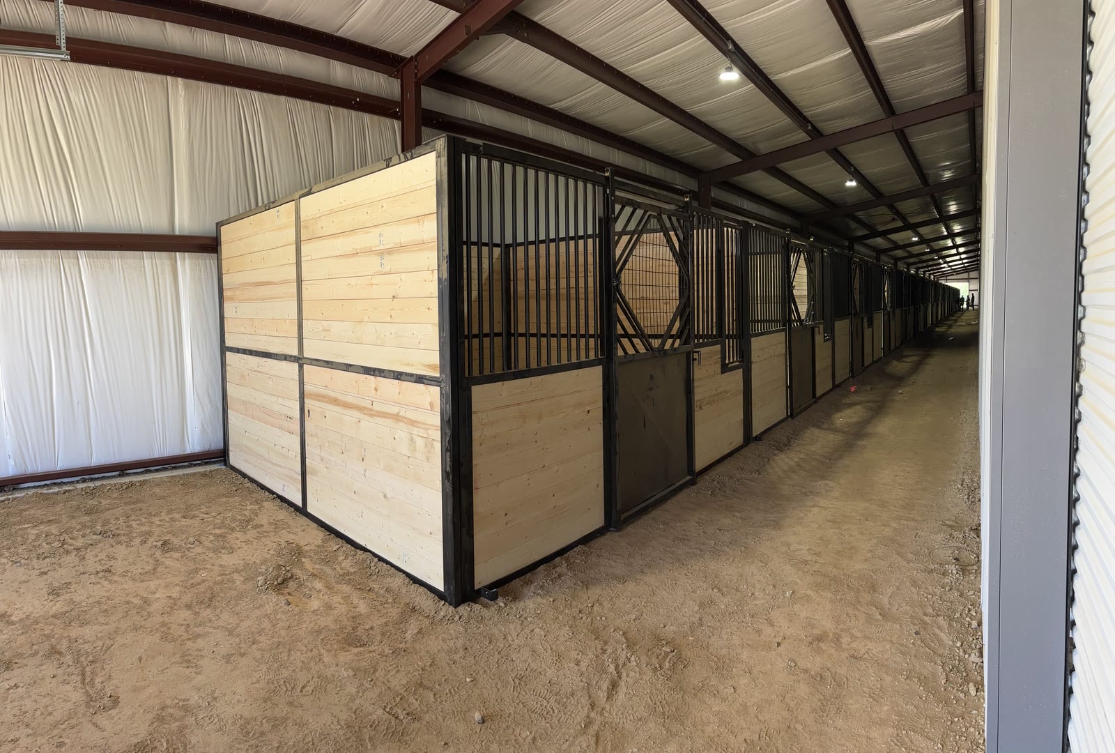 End of the finished stall run at Flying F Arena in Harrisburg, Arkansas: black steel frames with light wood infill, barred stall fronts running down the aisle